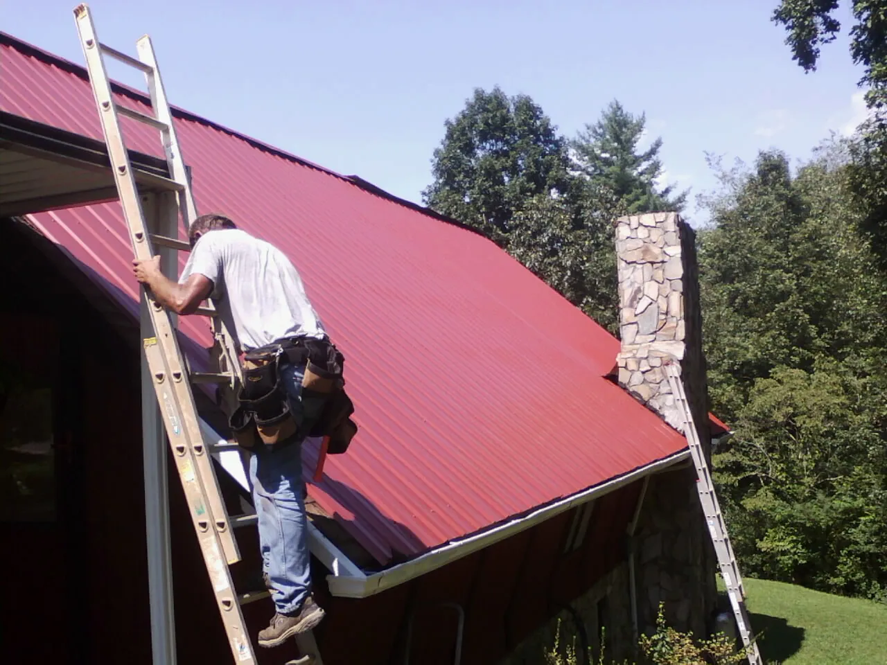 Roofer on a ladder beside a red metal roof and stone chimney — Soffit & Fascia Repair by Roofing Brothers in Edgewood